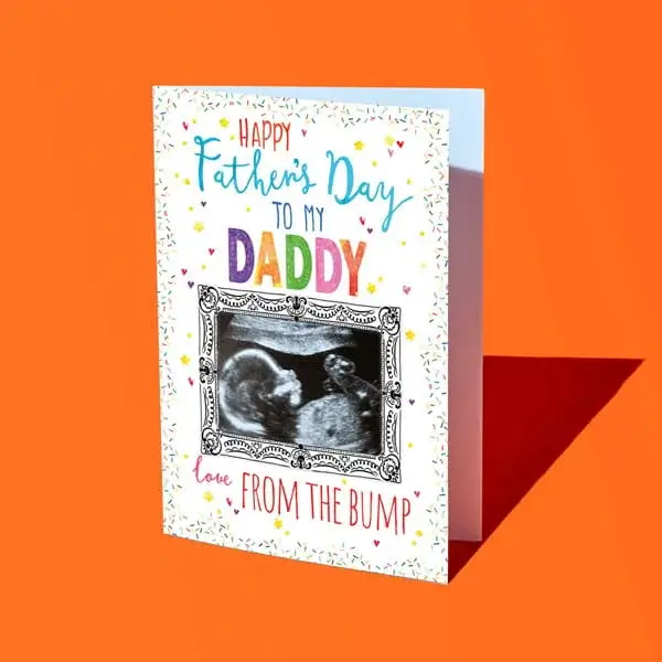 A Fathers Day card featuring a baby scan photo framed with decorative detail, colourful confetti accents, and text reading Happy Father’s Day to my Daddy love from the bump, displayed against a bright orange background.